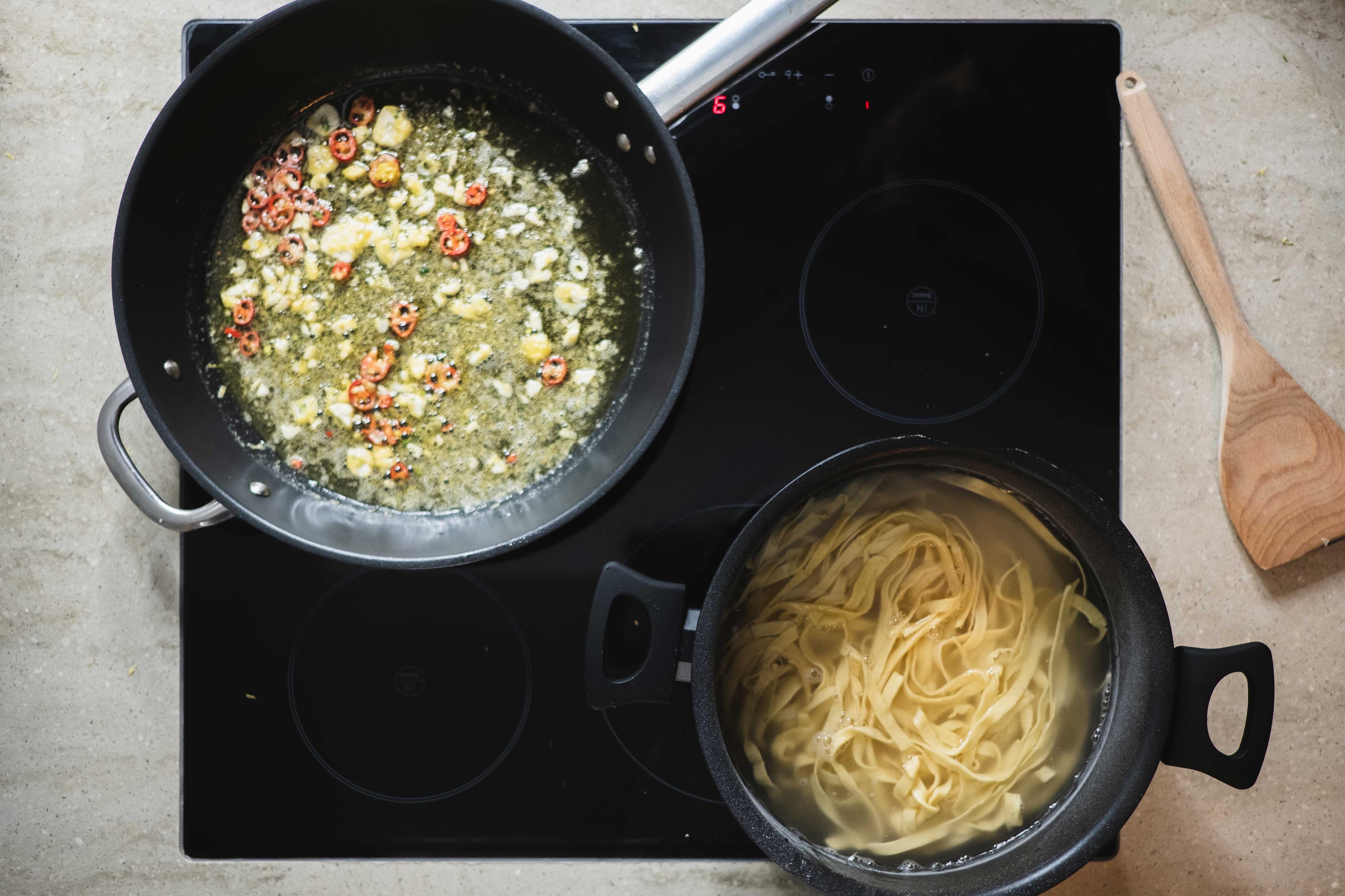 Chili, Zitronenabrieb und grob gehackter Knoblauch werden in einer Pfanne in Butter und Öl angebraten. Daneben Nudelwasser.