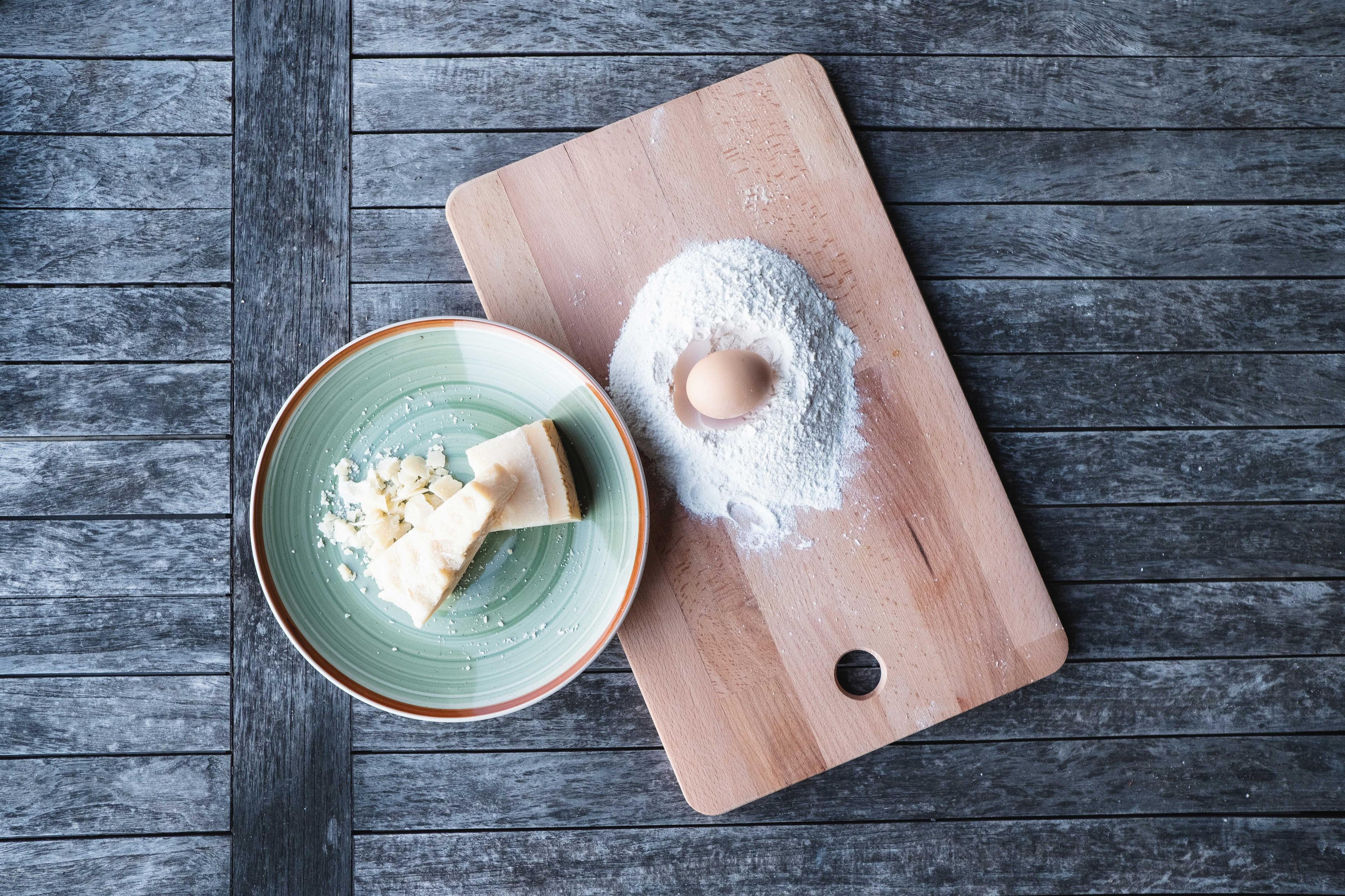 Brett mit Mehl in dessen Mitte in einer Kuhle ein Ei liegt, daneben dekorativ ein Parmesan in einer grünen Schüssel